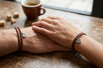 Deux poignets portant des bracelets de couple en cuir tressé avec breloque cœur, mains enlacées sur une table de café en bois